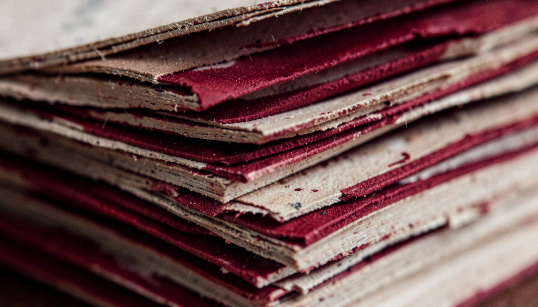 Extreme close-up photograph of stacked weathered pages and ink-stained edges in muted crimson and sepia tones, evoking an old keepsake archive.
